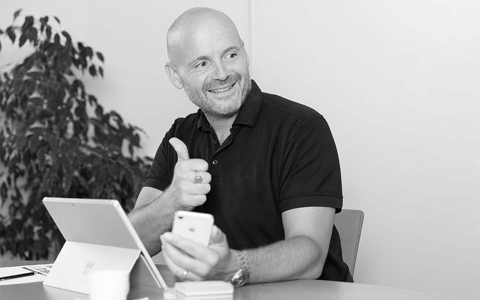 Ein lächelnder glatzköpfiger Mann in einem dunklen Polohemd sitzt an einem Schreibtisch mit einem Tablet und einem Telefon und zeigt den Daumen nach oben - ein Mitarbeiter einer Werbeagentur. Im Hintergrund steht eine Topfpflanze. Das Bild ist in schwarz-weiß gehalten.