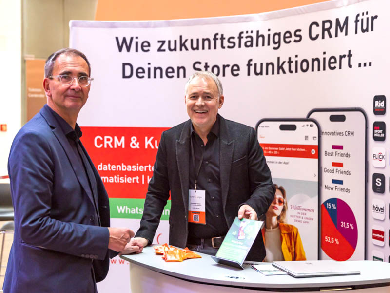 Zwei Männer stehen an einem Stand mit einer Ausstellung über CRM für Geschäfte. Einer hält eine Broschüre in der Hand, beide lächeln. Hinter ihnen ist ein Banner mit Smartphone-Bildschirmen und einem deutschen Text über zukunftssichere CRM-Lösungen zu sehen.