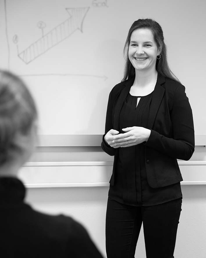 Eine Frau in Business-Kleidung steht lächelnd da, während sie einer anderen Person etwas präsentiert; auf dem Whiteboard hinter ihr ist ein Diagramm zu sehen, das einen Aufwärtstrend zeigt – eine dynamische Szene, die die professionelle Energie einer modernen Werbeagentur einfängt.