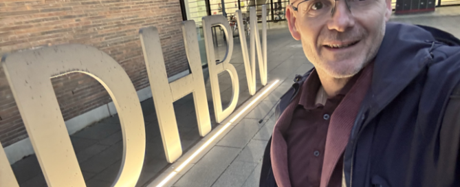 Ein lächelnder Mann mit Brille und Jacke macht ein Masterclass Gamification-Selfie vor großen, beleuchteten Buchstaben, die DHBW bilden.